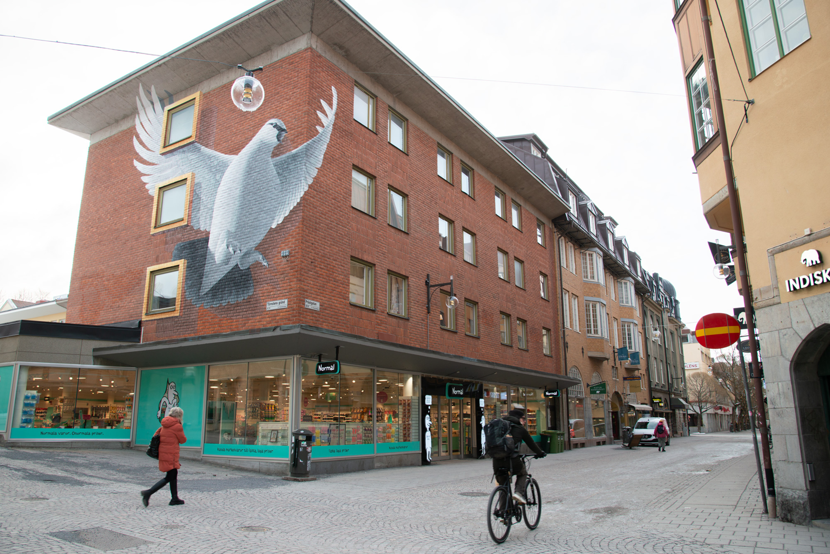 Byggnad i tegel med 4 våningar. På kortsidan är det en stor fasadmålning på en fågel. Butiker i ned