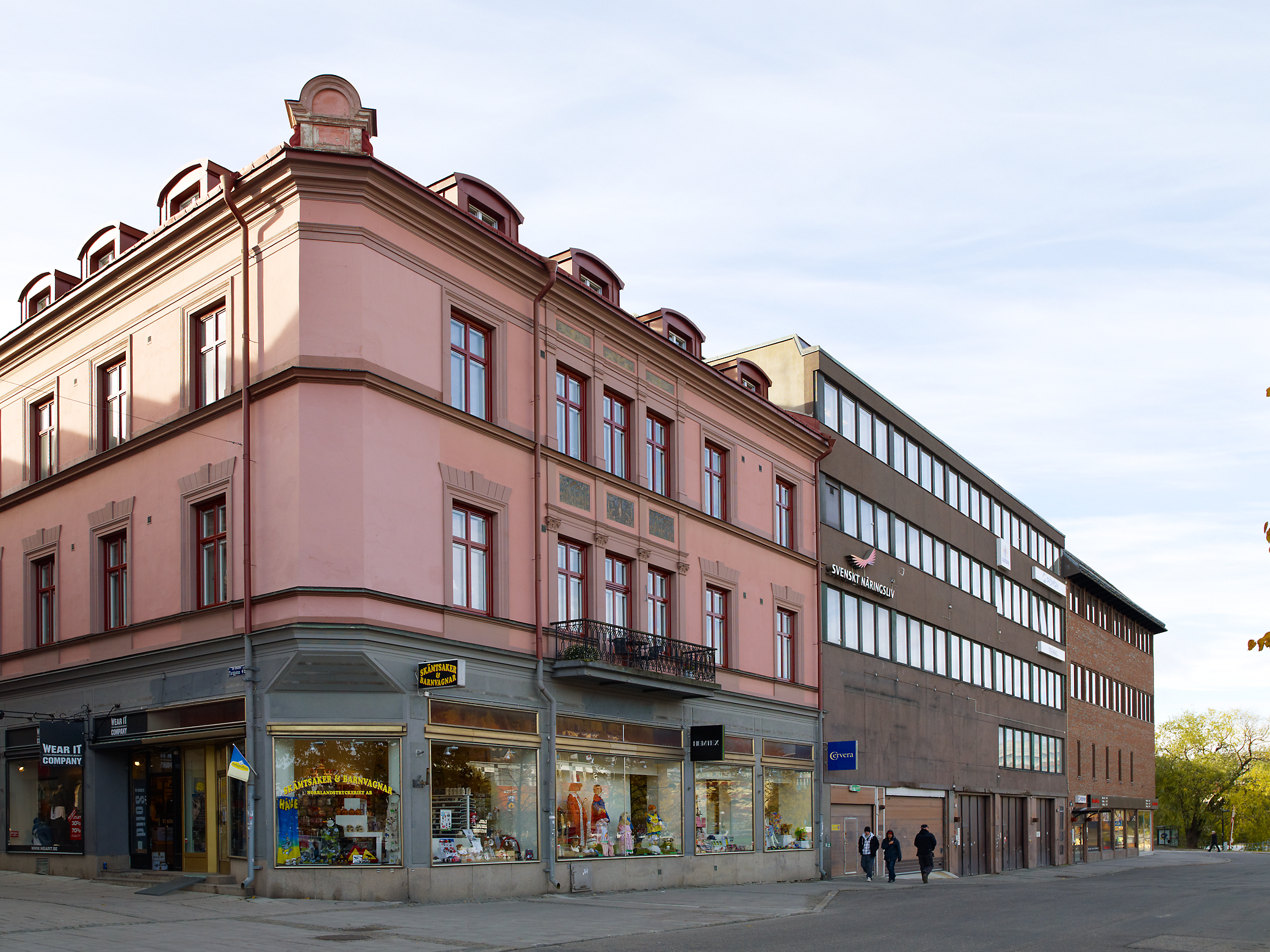 3 stycken sammansatta byggnader. I hörnet en rosa byggnad i 3 våningar med 1 balkong. Mittersta hus